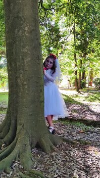 A girl in a white dress and a veil, the image of a scary bride, moves like a zombie on the street, in the park on Halloween
