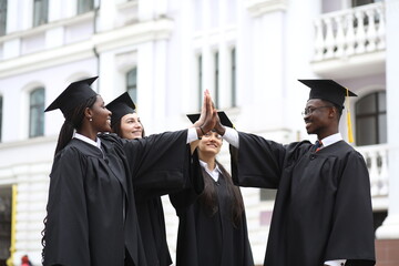 Portrait of a group of smiling happy multiracial international graduates students hugging and having fun in a university graduate gown and holding diploma outdoor. Education and graduation concept.