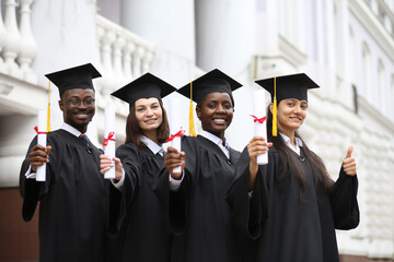 Portrait of a group of smiling happy multiracial international graduates students hugging and having fun in a university graduate gown and holding diploma outdoor. Education and graduation concept.