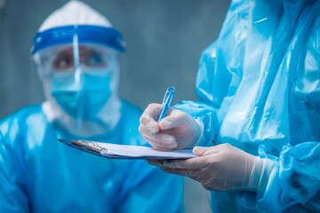 Two medical professionals in blue protective suits are focused on patient care. One records notes while the other observes attentively, emphasizing teamwork and diligence in a challenging environment