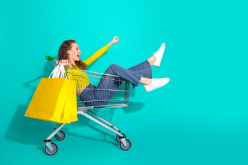 Excited young girl sitting in a shopping cart with yellow bags against a teal background, showcasing casual style and lively charm © deagreez