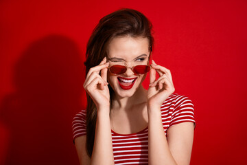 Attractive young woman with charming smile posing in red sunglasses on vibrant red background wearing striped t-shirt