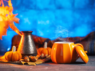 Hot coffee in an orange pumpkin cup. Autumn leaves and a warm knitted scarf. Blue background.