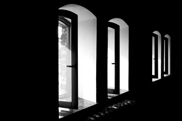 Black and white photo of arched windows with open frames in a dark interior. Minimalist architectural composition with strong contrast of light and shadow.