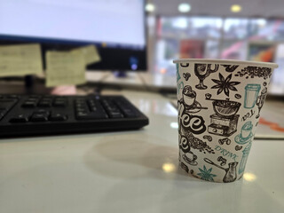 Paper coffee cup on office desk with blurred computer screen and keyboard in the background. Casual work environment with soft natural lighting.
