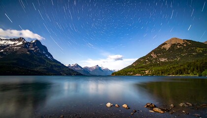 Obraz premium A long exposure shot revealing star trails above a calm lake and mountain landscape at night