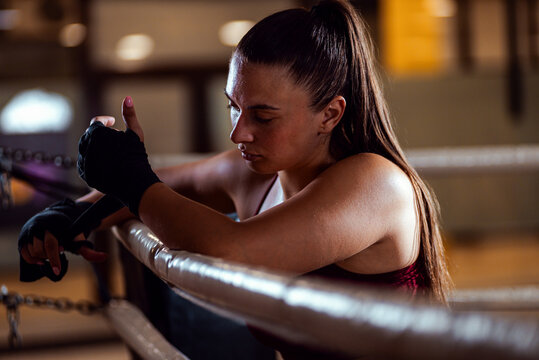 Female boxer leaning against the ropes of a boxing ring wrapping hands with boxing strap. - Powered by Adobe