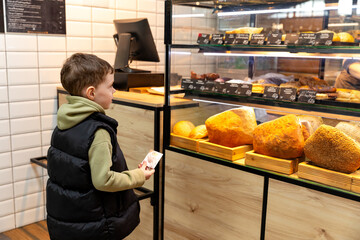 Cute child little boy choosing baked goods or pastry in bakery shop. Little boy buying bread or buns at grocery supermarket and paying by credit card. High quality photo
