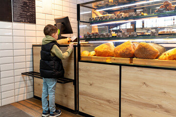 Cute child little boy choosing baked goods or pastry in bakery shop. Little boy buying bread or buns at grocery supermarket and paying by credit card. High quality photo