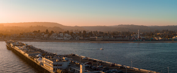 Obraz premium Aerial view of Santa Cruz, California, featuring the Giant Dipper rollercoaster, a long pier with parked cars, calm waters, and a serene sunset.