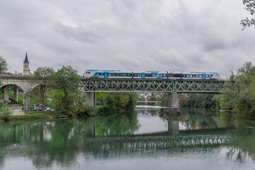 A modern blue passenger train crosses a steel railway bridge over the Krka River in Novo mesto, Slovenia. The church tower, lush trees, and river reflections create a peaceful scenic view.