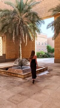Riyadh, Saudi Arabia, 17.05.2025. Al Murabba Historical Palace. Blonde woman in black long dress walking near fountain with palm trees. Vertical video.