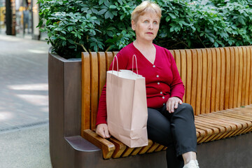 An elderly woman with a shopping bag is sitting in a shopping mall.
