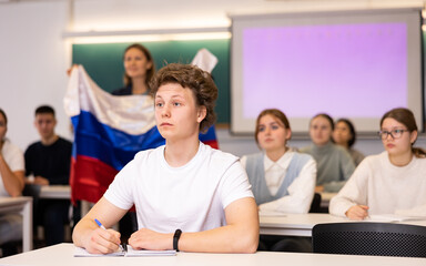 Students sit in classroom, teacher stands behind them with Russian flag