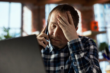 Distraught employee in home office sitting at desk in front of laptop, answering phone call, receiving bad news. Businessperson using notebook to do job online, receiving worrisome telephone call
