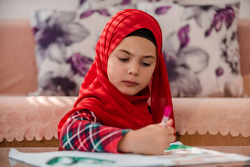 A little girl in hijab writing or painting in a notebook at a home.