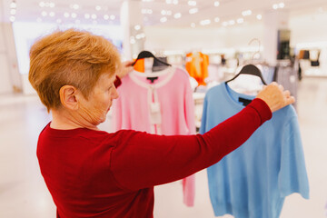 An elderly woman chooses between a T-shirt and a sweater in a store.