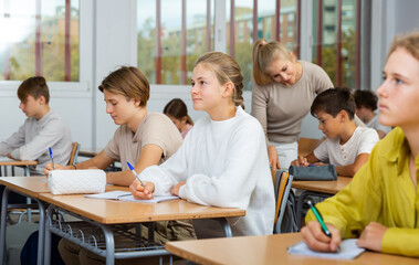 Teen boys and girls sitting at desk in classroom full of pupils during lesson