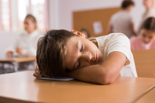 Portrait of tired bored school boy lying and sleeping at desk in classroom during lesson - Powered by Adobe