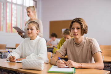 Fototapeta premium Teenage girl and boy sitting together at desk and doing tasks in classroom..