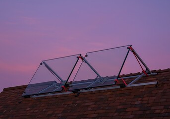 Fototapeta premium On the red brick roof a metal frame holds paired Glass glas panels secured with rubber straps and soft pads. The temporary setup suggests rooftop work in progress under a purple sky.