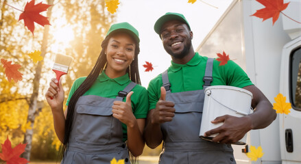 Professional African American painters team smiling with thumbs up. Man and woman in uniform with paint and brush outdoors. Home improvement and renovation service in autumn