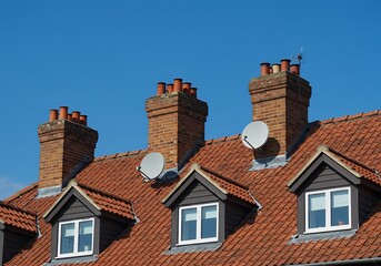 Two prominent satellite dishes sit atop a sloped roof with gabled windows under a clear, bright blue sky. The roof features classic red tiles and brick chimney structures.