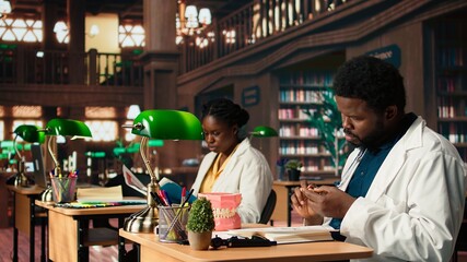 Medical student practices dentistry techniques with a plastic jaw model in a classic library, doing physical research for stomatology thesis. Developing professional skills for the career. Camera A.