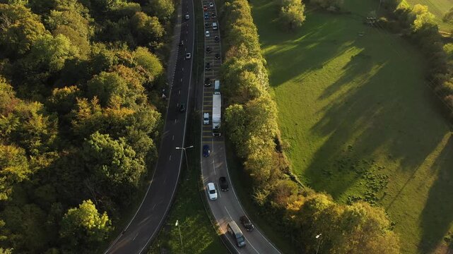 An aerial shot focusing on a road segment where a lane from a side road merges with the main dual carriageway, illustrating the flow of traffic with a long queue in one direction, framed by trees and 