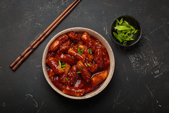 Stir fried rice cakes tteokbokki simmered in spicy sauce, garnished with green onions and sesame seeds, served on a black background with chopsticks top view. Popular dish of Korea