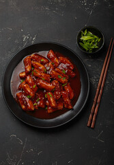 Stir fried rice cakes tteokbokki simmered in spicy sauce, garnished with green onions and sesame seeds, served on a black background with chopsticks top view. Popular dish of Korea