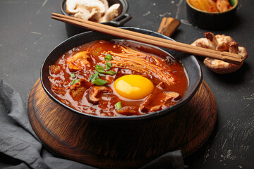 Top view of spicy kimchi jjigae soup in a black bowl with egg yolk, enoki, shiitake, tofu and green onions, with chopsticks and cooking ingredients, cuisine of Korea