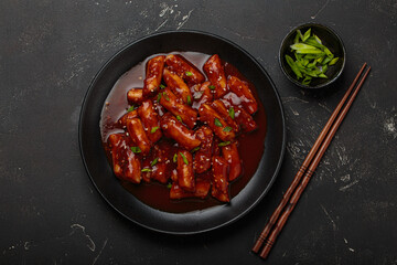 Stir fried rice cakes tteokbokki simmered in spicy sauce, garnished with green onions and sesame seeds, served on a black background with chopsticks top view. Popular dish of Korea