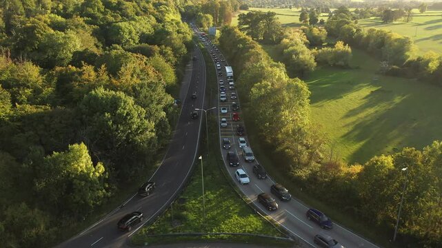 A wide aerial view of a dual carriageway passing through a valley, showing one side heavily congested with slow-moving traffic, contrasted by the clear lanes on the opposite side, surrounded by lush, 