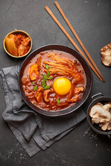 Top view of spicy kimchi jjigae soup in a black bowl with egg yolk, enoki, shiitake, tofu and green onions, with chopsticks and cooking ingredients, cuisine of Korea