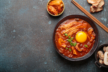 Top view of spicy kimchi jjigae soup in a black bowl with egg yolk, enoki, shiitake, tofu and green onions, with chopsticks and cooking ingredients, cuisine of Korea, copy space