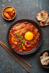 Top view of spicy kimchi jjigae soup in a black bowl with egg yolk, enoki, shiitake, tofu and green onions, with chopsticks and cooking ingredients, cuisine of Korea