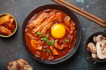 Top view of spicy kimchi jjigae soup in a black bowl with egg yolk, enoki, shiitake, tofu and green onions, with chopsticks and cooking ingredients, cuisine of Korea