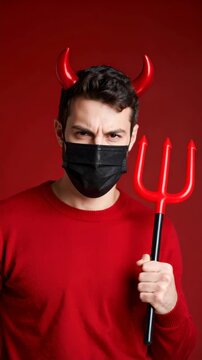 A man in a black medical mask and devil horns holds a trident against a red backdrop for Halloween