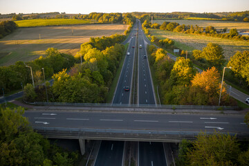 A high-angle aerial view of a dual carriageway motorway passing underneath an overpass, flanked by lush green trees and open farmland bathed in the warm light of the setting sun.