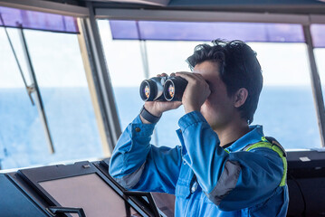 Obraz premium A male deck officer on the bridge of a merchant ship is looking through binoculars. 
