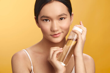 Asian woman holds a clear bottle near her face, presenting a skincare moment with bright studio...