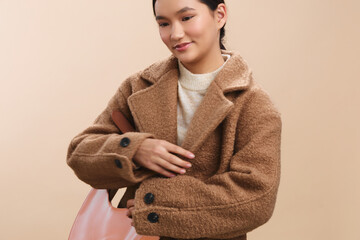 asian woman wearing a warm brown coat stands against a neutral backdrop, presenting contemporary...