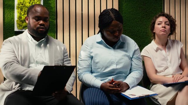 Anxious diverse job seekers awaiting job interview discussion with manager, showcasing anticipation in office hallway. People preparing with information for the recruitment process. Camera B.