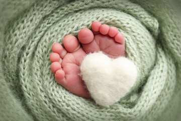 The tiny foot of a newborn baby. Soft feet of a new born in a olive green wool blanket. Close up of toes, heels and feet of a newborn. Knitted white heart in the legs of a baby. Macro photography.