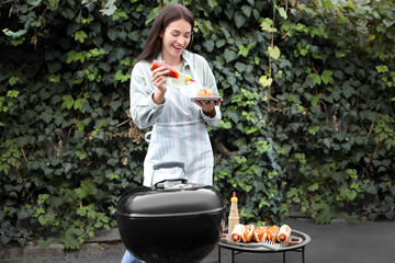 Young happy woman adding ketchup onto tasty hot dogs near barbecue grill outdoors