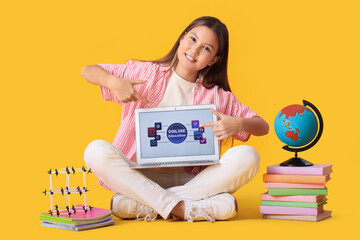 Happy teenage Asian girl with school stuff pointing at laptop on yellow background. Online education concept