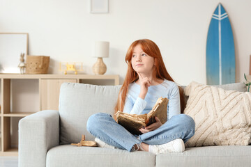 Fototapeta premium Cute teenage thoughtful redhead girl reading adventure book on sofa at home