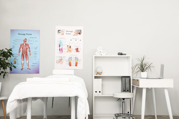 Interior of massage therapist's office with workplace, couch, shelving unit and posters on wall in modern medical centre