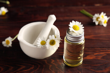 Bottle of chamomile cosmetic oil and mortar with flowers on wooden background, closeup